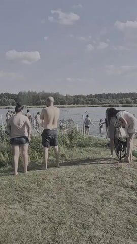 The video shows a crowded beach on a sunny day. People are swimming in the water, sunbathing on the sand, and interacting with each other. There are trees and bushes in the background, and the sky is blue with some clouds. Several inflatable objects, such as a green alligator float and a black inner tube, are visible on the beach and in the water. Two women are standing near the water's edge, one of whom is holding a white bag. A man is standing in the foreground facing away from the camera, wearing black swim trunks. Other individuals are scattered across the beach and in the water.