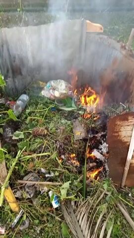 The video shows a pile of trash and vegetation burning in an outdoor enclosure made of corrugated metal and wood. Flames are visible in the center of the pile, and smoke is rising. The trash includes plastic bottles, food packaging, and what appear to be bags of garbage. The vegetation is a mix of green leaves and dried grass. The overall scene suggests an uncontrolled fire in a waste disposal area.