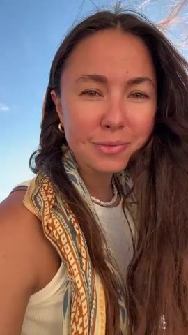 A young woman with long brown hair smiles at the camera. She is wearing a white tank top and a patterned scarf around her neck. The sun is shining brightly behind her, casting a lens flare across the image. The background shows a body of water and a blue sky with some clouds. The woman appears to be on a boat or a similar vessel, as there is a railing visible in the lower right corner of the frame. Her hair is blowing in the wind.