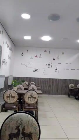 The video shows the interior of a restaurant. The walls are decorated with various pictures and text. There are tables and chairs arranged throughout the space. In the foreground, there are several empty tables and chairs. Further back, two men are seated at a table, eating. The overall atmosphere appears to be calm and quiet.