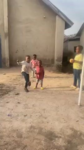 In a wide, full shot, four young children joyfully dance and frolic in a sandy courtyard. The child on the left wears a white t-shirt with a print and dark pants. The second child has on a red-and-white checkered shirt. The third child from the left sports a bright yellow shirt, while the child on the right is clad in a red tank top. All the children are barefoot, except for the child on the right who wears yellow sandals. They exhibit enthusiasm and appear to be in good spirits.

The courtyard is enclosed by two buildings with a simple, grey facade. The building on the left has a visible blue door. A white pole stands on the right edge of the frame. The ground is sandy and uneven. The sky is visible at the top of the frame. The scene is brightly lit, indicating daytime.