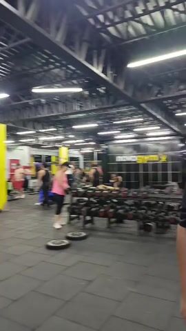 The video depicts the interior of a gym with several people working out. The gym has a high ceiling with exposed metal beams and rows of fluorescent lights. The floor is covered with black rubber tiles. There are various exercise machines and weightlifting equipment scattered throughout the space, including dumbbells, barbells, and weight plates. Several individuals can be seen performing exercises, while others are walking around or resting. A large advertisement banner with the word "TECHNOFIT" in bold vertical letters is visible, as is some artwork on the walls. The camera appears to be moving.