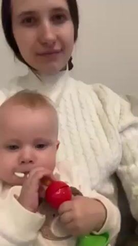 A woman holding a baby in her lap drinks from a coffee cup. The woman is wearing a white sweater. She has dark hair and fair skin. The baby is wearing a white outfit with a small teddy bear print. The baby has blond hair and light blue eyes. The baby is holding a red and green rattle.  The background is a plain white wall. Music can be heard playing.