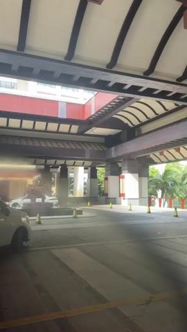 A doorman in a white uniform stands to the right of the entrance to a building. Yellow cones are placed on the ground. The video pans to the left, showing an indoor area with a skylight. Two people, one in a white shirt and shorts, the other in a white shirt and dark shorts, are seen near luggage carts. The video continues to pan, revealing more of the interior, with decorative horse statues and a large floral-like wall ornament. The video pans again, focusing on one of the horse statues.