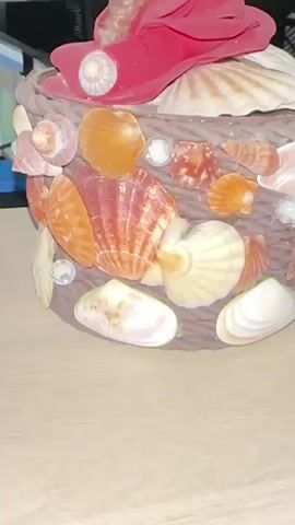 A close-up, slightly shaky, medium shot focuses on a decorative cylindrical object covered in various seashells and embellishments. The object appears to be made of a dark brown, woven material, possibly rope or twine. Numerous seashells of different shapes, sizes, and colors, including scallop shells, clam shells, and smaller spiral shells, are attached to the surface. Some of the shells are light brown, orange, pink, and white. Several clear, faceted beads or rhinestones are also adhered to the object, adding sparkle. At the top, a large pink or fuchsia flower-like embellishment, possibly made of fabric or felt, is visible, along with some white shell pieces. The object sits on a light-colored, textured surface, likely a wooden table. The background is dark and out of focus, with a hint of blue and black visible on the left side. The lighting is somewhat dim, creating soft shadows. The video appears to be a short, static shot, possibly a close-up of a handicraft item.