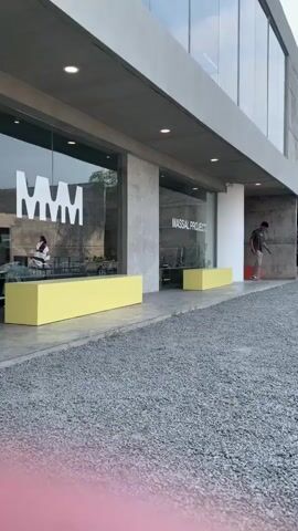 A man walks across a gravel area towards a modern building with large windows. A yellow bench sits in front of the building. Another man walks past a sign that says "SALE 50%". Several outdoor tables and chairs are visible in the gravel area.