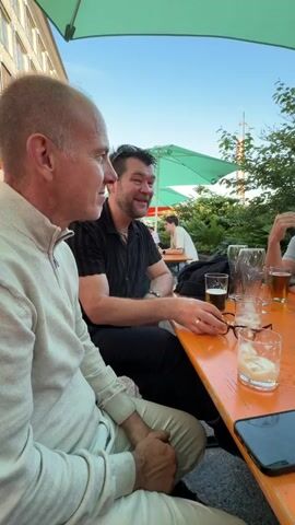 The video shows a group of people sitting at an outdoor table, likely at a restaurant or beer garden. An orange umbrella provides shade. There are several glasses on the table, some containing drinks, and a pack of cigarettes. A smartphone lies on the table in the foreground. The people are engaged in conversation, with some appearing to be laughing or gesturing. The setting is outdoors with greenery visible in the background. The lighting suggests it is late afternoon or early evening.