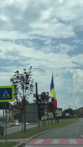 The video shows a Romanian flag flying high above a street on a cloudy day. The flag is a tricolor of blue, yellow, and red. The flag is mounted on a tall pole and is visible from a distance. The street is lined with trees and signs. There are cars driving on the street. The sky is filled with white clouds.