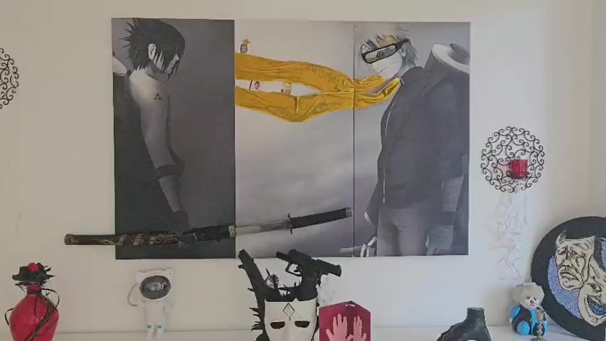 The video shows a wall display with several items. Dominating the wall is a triptych painting depicting two anime characters, one on the left and one on the right, with a yellow fabric flowing between them in the center. Below the painting, a sword is mounted horizontally. To the left of the painting is a red vase with black floral accents. To the right of the painting, there are two decorative wall sconces. Below the painting, on a white shelf, there are various objects including a white astronaut-like figure, a white mask with a diamond symbol and black embellishments, a red lantern with handprints, a black hand sculpture, and a small clown figure. To the far right, there is a round decorative plate with a depiction of two faces. The lighting suggests an indoor environment.