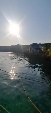 The video shows a bright sun in the sky over a body of water. In the distance, there is a shoreline with a building and trees. There is also a boat visible in the water. The water appears to be clear and calm. There is a fishing line visible in the foreground of the video.