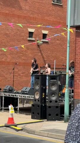 A short video clip shows an outdoor music performance with a brick building in the background. The building is decorated with colorful triangular flags strung across the upper portion of the wall. On a raised stage, three individuals are visible. The person on the left, a male, is playing a white electric guitar and singing into a microphone. He is wearing a black t-shirt and blue jeans. The person in the center, a female, is also singing into a microphone, wearing a light-colored top and dark shorts. To the right, a third person, whose gender is unclear, is playing a guitar. Prominently placed in front of the stage are large black speaker cabinets stacked on top of each other. A red and white traffic cone with a yellow warning sign is situated on the left side of the foreground, near the edge of a paved area marked with yellow lines. The overall lighting suggests it is daytime.