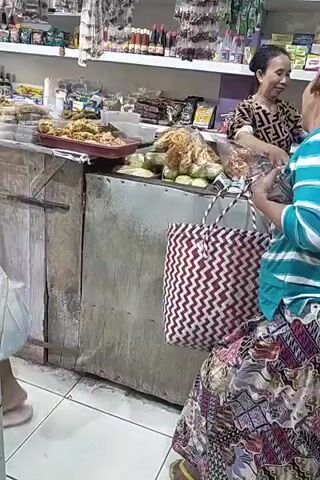 Two women are at a market stall, appearing to be interacting. One woman, facing away from the camera, wears a blue and white striped long-sleeved shirt and a patterned skirt, carrying a woven bag with a red and white chevron pattern. She has her hair pulled back in a bun. The other woman, partially visible, wears a patterned shirt and is smiling. The stall is filled with various food items and packaged goods displayed on shelves and in trays. The environment appears to be an indoor market or shop.