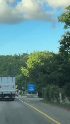 A first-person perspective video shows a view from inside a vehicle traveling on a road. The road is paved and has a yellow line marking the edge. A white truck with a gray trailer is in front of the vehicle. To the right of the road, there is a pedestrian walkway with a metal guardrail. Several trees and lush green vegetation cover the surrounding hills. In the distance, a blue billboard with Russian text and a logo is visible. A few people are walking on the pedestrian walkway. The sky is blue with scattered clouds. The video captures the movement of the vehicle as it drives down the road, passing by the truck, walkway, and billboard.