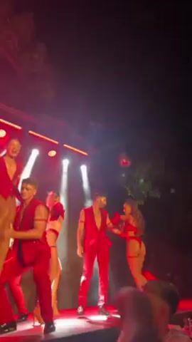 Here is a detailed description of the video:

The video captures a live stage performance, with a group of dancers dressed in red costumes. The men wear red vests over bare chests and red pants, while the women are in matching red lingerie-style outfits. The stage is lit with bright, colorful lights, creating a vibrant atmosphere.

The performance involves synchronized dance moves, with the dancers moving in unison to the music. In the background, a logo appears, suggesting a brand or event affiliation. The camera is hand-held and positioned in the audience, so only parts of audience members’ heads are visible in the foreground. 

Overall, the video depicts a high-energy dance performance, likely part of a stage show.