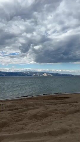 The video shows a view of a lake and mountains from a beach. The foreground is sandy beach with ripples. The lake water is a dark grayish-blue with small waves. In the distance, there are mountains with some snow visible on the peaks. Above, the sky is filled with thick, gray clouds with patches of blue sky peeking through. The camera zooms in slightly on the mountains. There are no people or other discernible objects in the shot.