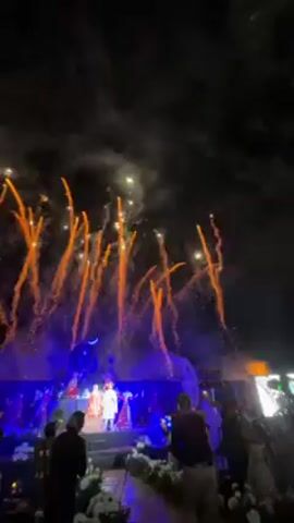 The video captures a nighttime celebration, possibly a wedding, with fireworks illuminating the sky. A stage is visible, lit with blue lighting, where a couple dressed in what appears to be traditional Indian attire stands. Guests are present, some of whom are taking photos with their phones. The fireworks are prominent, displaying bright orange and white trails against the dark sky. The overall atmosphere is celebratory and festive.