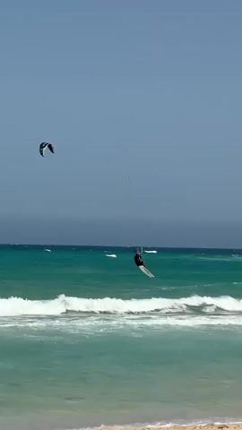 The video shows a beach scene with kite surfers in the turquoise water. The camera is positioned on the sand, looking out towards the ocean. In the foreground, there is a sandy beach. In the middle ground, there are white waves breaking. In the background, there are multiple kite surfers riding their boards and being pulled by kites. One kite surfer is in the foreground, closer to the camera, and appears to be jumping or performing a trick. Further out in the water, other kite surfers are visible, some closer together and some further apart. The sky is blue with some darker clouds on the horizon. There is a seagull on the beach in the bottom left corner of the frame.