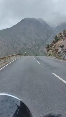 A first-person point-of-view video captures a motorcycle traveling on a wet asphalt road that winds through a mountainous landscape under a cloudy sky. The road has a yellow guardrail on the left side, and a sheer rock face on the right. The mountains are rugged and gray, with patches of snow visible on their upper slopes. The camera is positioned low, showing the front of the motorcycle, including a black reflective surface, possibly a headlight or part of the fairing, with water droplets on it. A white line and a double white line mark the lanes on the road. In the distance, there are a few green signs and a small light pole. The overall atmosphere is cool and damp, suggesting recent rain or fog.