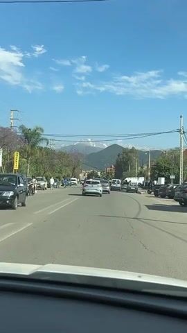 The video shows a street scene in a town, possibly in Morocco, during a sunny day. The road is lined with trees and buildings.  Traffic is moderate, with cars including a grey Renault Clio visible. In the background, there are mountains with snow-capped peaks. People are walking on the sidewalks. The sky is blue with some clouds. The camera is likely mounted in a vehicle, providing a driver's perspective.