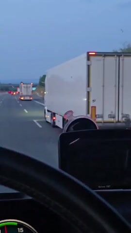 The video shows a view from inside a vehicle on a highway at dusk. The camera is positioned behind a steering wheel and a dashboard device. In front of the vehicle, a white semi-trailer truck is in the right lane, its brake lights illuminated. Further ahead, another white semi-trailer truck is visible in the left lane, also with illuminated brake lights. The sky is a deep blue, indicating the transition from day to night, and there are trees and vegetation visible on the side of the highway. The road appears to be a multi-lane highway.