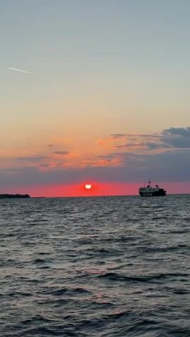 The video shows a stunning sunset over the sea. The sun is a vibrant red, partially obscured by clouds on the horizon. A cargo ship is visible in silhouette, floating on the calm water to the right. A faint contrail from an airplane stretches across the clear upper sky. An islet or land mass is visible far on the left horizon. The waves on the water are gentle, and the scene is peaceful.