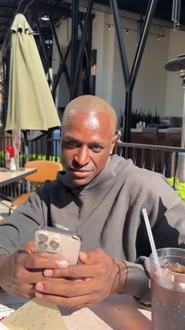 A man is sitting at an outdoor restaurant. He is wearing a gray hoodie, has blonde hair and is holding a phone. There is a drink with a straw in front of him. The camera zooms in as he sends a kiss towards the camera. He then licks his lips.