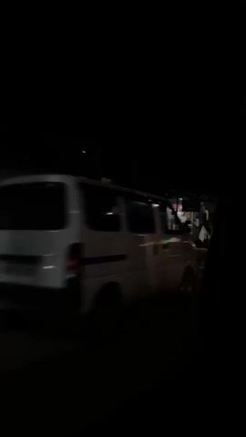 The video shows a view from inside a vehicle at night, looking out at a street lined with shops and buildings. The lighting is dim, with some artificial lights illuminating storefronts and the street. Several shops appear to be open, with goods displayed outside. The camera pans across the scene, providing glimpses of different businesses and structures. There are no clear views of individuals or specific actions taking place. The environment appears to be a commercial area at night.