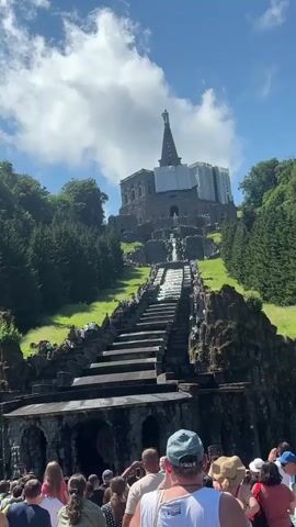 A vertical video captures a wide shot of the Bergpark Wilhelmshöhe in Kassel, Germany. The scene focuses on the Hercules monument and the cascading water features leading up to it. The monument, a large stone structure topped with a gilded statue of Hercules, is partially covered in scaffolding. Below it, a grand staircase with water flowing down its center stretches towards the viewer. The stairs are lined with people on both sides, many of whom are looking up towards the monument. The surrounding landscape is lush green, with dense evergreen trees on either side of the monument and a bright blue sky with fluffy white clouds overhead. In the foreground, the backs of a crowd of people are visible, all facing the monument and appearing to be tourists. The camera is positioned low, looking upwards, emphasizing the scale of the monument and the park. The video has a slight shakiness, indicating it was likely recorded handheld.
