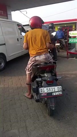 A person wearing a red helmet and an orange shirt sits on a motorcycle at a gas station. The motorcycle is black with a license plate that reads AG 5831 DO. In the background, there is a white van, another motorcycle, and a gas pump with signs that say PERTALITE and PERTAMINA DEX. The ground is paved with bricks. The scene appears to be outdoors under a covered area.