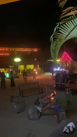 The video shows a lively nighttime scene, likely at a beach bar or resort, with multiple people visible. In the foreground, there's a small bonfire surrounded by what appears to be tree stump seats. People are sitting and standing around, some enjoying the fire. In the middle ground, individuals are performing with fire torches, spinning and moving them in a show. Several spectators are watching. The background features a building adorned with lights, including a prominent "Chang" beer sign. Palm trees add to the tropical atmosphere. The overall impression is a relaxed and festive gathering.