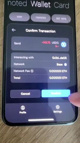The video shows a close-up of a smartphone screen displaying a cryptocurrency wallet application called "noted Wallet Card". The screen shows a "Confirm Transaction" page. The user is sending -148.75 USDC to an address identified as 0x3d_de05 on the "Base" network. The network fee and total transaction cost are both listed as 0.000001 ETH. A finger is seen pressing the "Confirm" button on the right side of the screen. The transaction is currently processing as indicated by a spinning icon on the "Confirm" button. The background of the phone is a wooden surface.