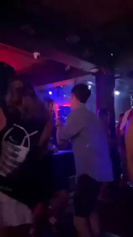The video shows a group of people dancing in a dimly lit nightclub. The lighting shifts between blue and red, creating a vibrant, energized atmosphere. Several individuals are visible, some holding drinks and moving to the music. The camera moves through the crowd, capturing the lively interactions and dancing motions of the people. Speakers and other club equipment can be seen in the background.