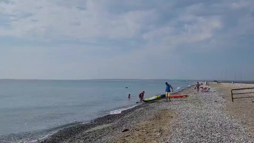 The video shows a wide shot of a pebble beach with the ocean stretching to the horizon under a cloudy sky. In the foreground, several people are present on the beach and in the shallow water. Two people are standing near the water's edge with what appear to be paddleboards. One person, wearing a blue shirt and yellow shorts, is interacting with a paddleboard. Another person in a red shirt is further down the beach. Several other individuals are visible in the water further out. The waves are gently lapping at the shore. The overall scene depicts a calm day at the beach with recreational activities taking place.