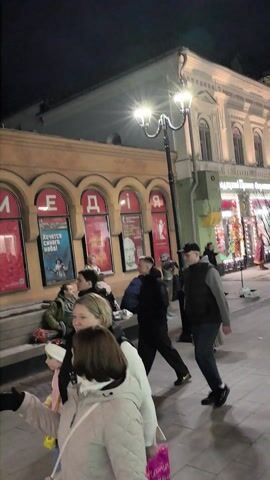 The video shows an outdoor scene at night. A group of people are walking down a pedestrian street, with buildings on either side. One building has arches displaying the word "KOMEDIA." There are many other people walking around, and the area appears well-lit by streetlights. Some of the buildings are decorated with lights.