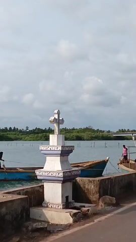 The video captures a stationary, medium-shot of a waterfront scene under a cloudy sky. The focus is on a white, ornate Christian cross mounted on a tiered pedestal, positioned on a concrete barrier. In the background, there is a body of water lined with lush greenery and a few distant structures, possibly including a bridge. Two small, blue wooden boats are moored along the waterfront. One boat is occupied by a person sitting near the front, while another individual stands in the second boat. The scene suggests a serene coastal or lakeside setting, potentially a place of worship or commemoration near a waterway used for fishing or transport.