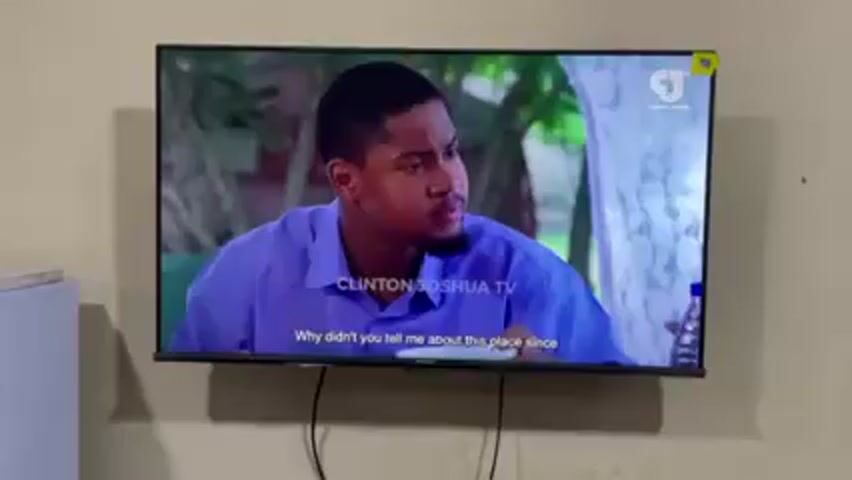 A wall-mounted television displays a scene of two men eating. The television screen also shows a logo for "Clinton Joshua TV" in the lower center of the screen. The men are close to the table. A bottle of water is on the table in front of one of the men.  The television is mounted on a plain wall. To the left of the TV is a white, rectangular object.