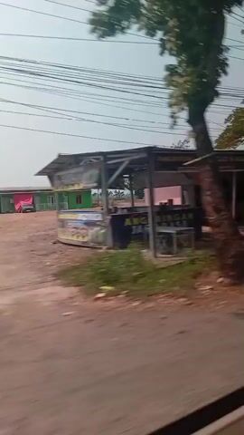 The video shows a side view of a road with a slightly overcast sky. The camera appears to be in motion, possibly from a vehicle, as the scenery is blurred and moving from left to right. On the right side of the road, there are various small buildings and structures, some appearing to be shops or homes with corrugated metal roofs. A scooter is parked in front of one of the buildings. There are also trees, overgrown vegetation, and power lines visible. The road surface appears to be dirt and gravel, with some drainage ditches along the side. The overall impression is of a rural or semi-rural area.