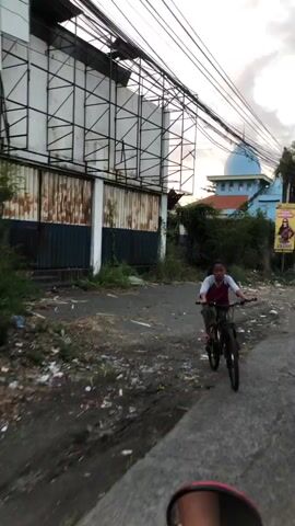A young girl rides a bicycle down a dirt path next to a busy road. She is wearing a maroon and white shirt and pink shoes. The path is lined with trash and debris. In the background, there is a large building with a metal frame and corrugated metal siding. There is also a blue dome visible in the distance. The sky is overcast.