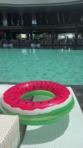 The video shows a poolside scene. In the foreground, a watermelon-shaped inflatable ring lies on a white lounge chair. The pool is to the left and center, with clear blue-green water. In the background, there is a building with multiple levels and large windows. People can be seen sitting at tables under a covered area near the pool. The lighting suggests it is daytime, likely sunny.