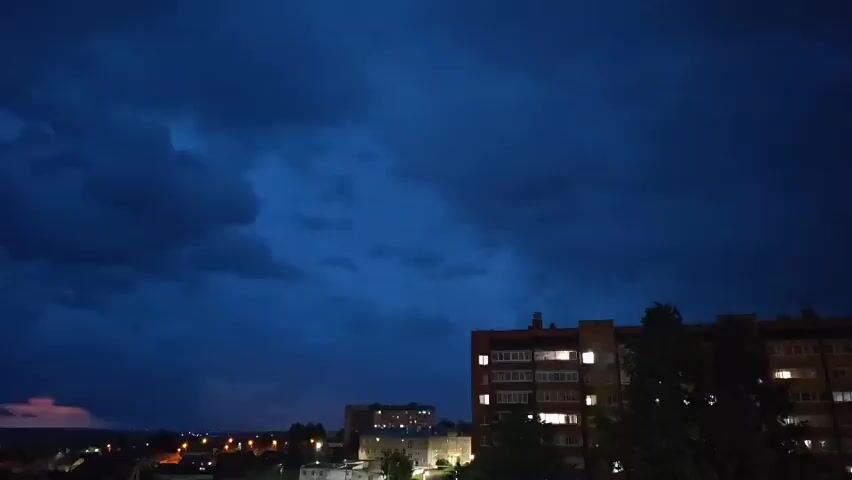 The video shows a nighttime cityscape under a dark, stormy sky. The clouds are heavy and appear to be in motion, with a dark blue hue. In the foreground, there are buildings, including a multi-story apartment building with lights on in some windows. Trees are also visible. In the distance, there are more buildings and lights, suggesting a town or city. There is a faint, reddish glow on the horizon, possibly from distant lightning or city lights. The overall atmosphere is dark and ominous.
