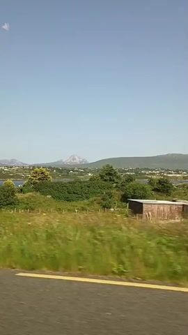 The video captures a view from a moving vehicle, likely a car, traveling along a road. The scenery outside the vehicle is a rural landscape. In the distance, mountains are visible under a clear blue sky. Closer to the foreground, there are green fields, trees, and some rural buildings. One prominent building is a white house with a dark roof, featuring a small green shed attached to its side. Further along, another white house with a textured finish and a chimney is seen. A blue car is parked in a gravel driveway next to one of the houses. The road has a yellow line painted on it, indicating it's a traveled path. The overall impression is a sunny day in a countryside setting.