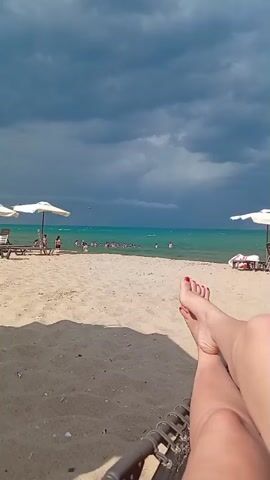 The video shows a first-person perspective of someone relaxing on a beach. In the foreground, the person's legs and feet are visible, with toenails painted red. They are lying on a beach chair. In the background, there is a sandy beach with several white umbrellas and lounge chairs. Further back, people are swimming and playing in the turquoise ocean. The sky is overcast with dark, dramatic clouds, suggesting an impending storm. A large blue umbrella is visible in the upper left corner of the frame.