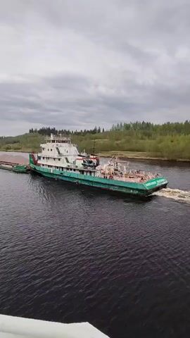 A large tugboat is traveling down a river on a cloudy day. The tugboat is teal and white and pulling a long barge. The river is dark and there is vegetation on either side. The video is taken from an elevated position, possibly a bridge. The tugboat is moving at a moderate speed, leaving a wake behind it.