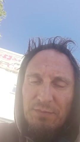 A man is facing the camera, speaking with his mouth open slightly. He has dark hair that is messy and stands on end. He has a thin beard and a gold tooth. He is wearing a gray hooded sweatshirt. Behind him, a white building with red letters is partially visible. Above the building, the sky is a clear blue.