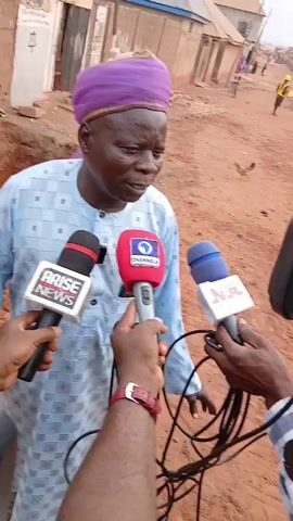 An individual wearing a light blue patterned shirt and a purple turban is speaking into three microphones held by three people. The microphones are from Arise News, Channels, and NTA News. The person being interviewed is standing on a reddish-brown dirt road. The background shows a building with graffiti, other buildings in the distance, and several people. The person holding the Arise News microphone is wearing a dark blue blazer and a watch. The person holding the Channels microphone is wearing a red watch and a light blue and white checkered shirt. The person holding the NTA News microphone is wearing a light blue shirt and has a silver ring on their finger. There is a ditch or excavated area beside the building with graffiti.