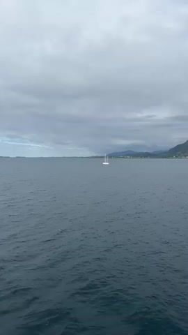 A sailboat with white sails is visible in the distance on a dark blue, choppy body of water. Beyond the sailboat, a coastal town with buildings and mountains can be seen. The sky is overcast with gray clouds. The camera then pans to the right, revealing the side of a white boat with black accents and a glimpse of the sea and the coastline in the background. Another, larger white boat with a dark stripe is sailing further out on the water. The video ends with a view of the ocean and the mountainous coastline under a cloudy sky.