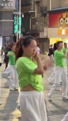 The video shows a group of Asian women practicing Tai Chi or a similar slow-motion exercise in an urban setting. They are all wearing matching lime-green shirts and white pants, performing synchronized movements to music. The setting appears to be a pedestrian area or plaza, with buildings and shops in the background with Chinese characters on their signs. Other people are seen walking by in the background, observing or passing through the area.