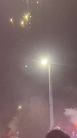 The video shows a group of people gathered at night under streetlights, holding a banner and lighting off fireworks. The banner is white and has writing on it in Cyrillic script. There is smoke in the air, likely from the fireworks, and some of the people are holding torches. The people seem to be enjoying themselves, celebrating, or protesting. The environment appears to be a street or public area, possibly during a celebration or demonstration. There are fireworks going off both in the sky and on the ground. One man is shown close to the camera with his back mostly to the viewer. His hair is short and dark. The camera seems to be handheld and moving slightly.