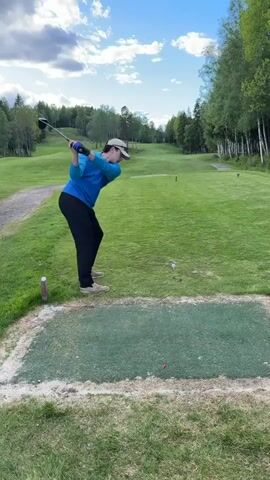 The video shows a man playing golf. He is standing on a green tee box with a golf club in his hands. He swings the club and hits a golf ball. The ball flies through the air towards the fairway. The background shows a golf course with green grass, trees, and a blue sky with clouds. Two other tee markers are visible in the distance. A dirt golf cart path is to the side of the fairway. A small, red, discarded golf tee is visible on the tee box.