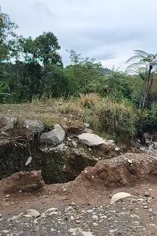The video shows an outdoor scene with a construction site and lush vegetation. A partially constructed concrete building is visible on the left, with a blue door. In front of the building, a large tree fern stands out amidst dense green foliage. The ground is rough and uneven, with loose soil, rocks, and some puddles of muddy water, suggesting recent rain or construction activity. A wooden utility pole is also present in the foreground. The sky appears overcast. The camera is held at an angle, giving a sense of perspective and depth to the scene. The video appears to be recorded from a moving perspective, possibly while walking.